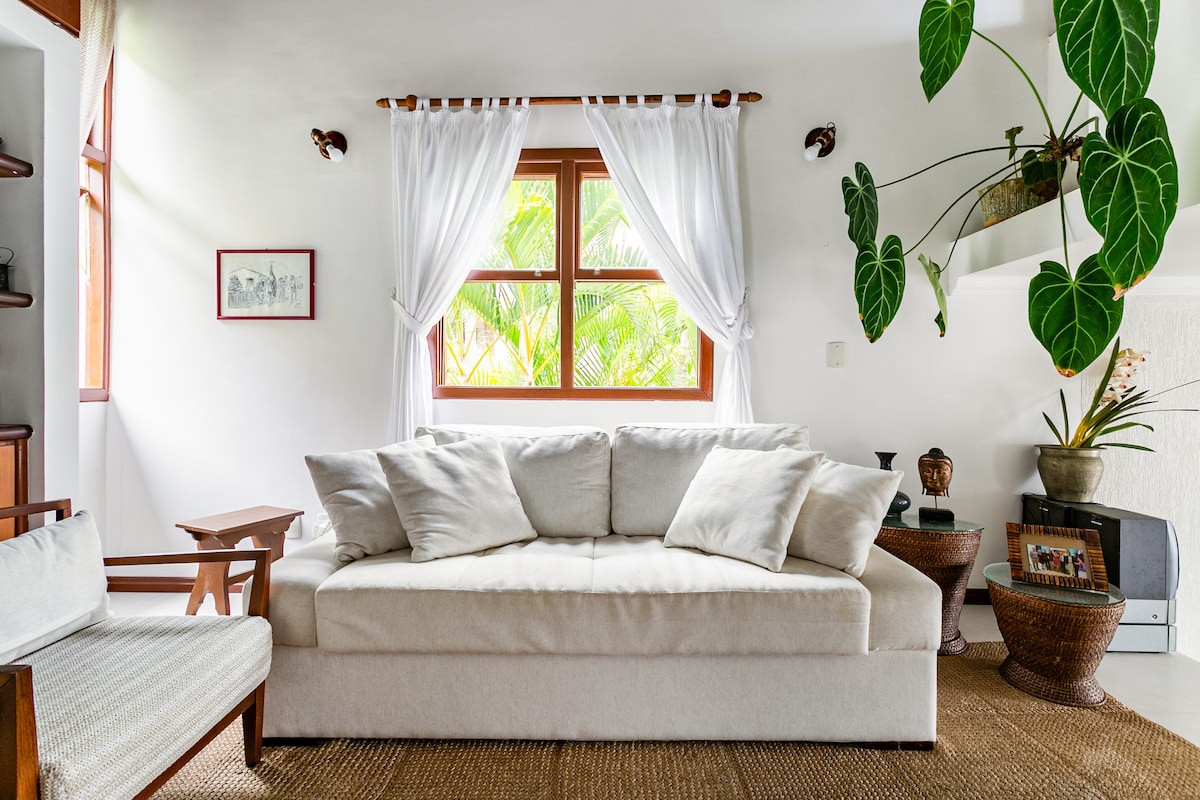 A bright living area features a large, white sofa with plush cushions, complemented by a wooden armchair. Natural light enters through a window adorned with sheer curtains, while green plants add a refreshing touch. Decorative elements are arranged on a shelf and side table.