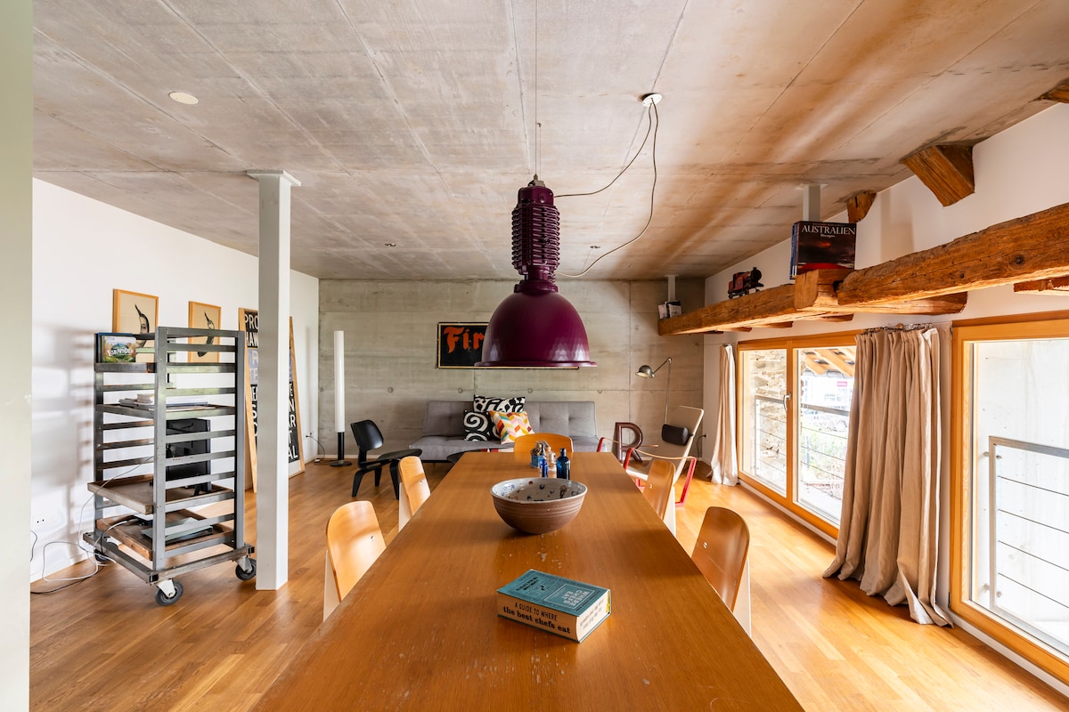 A spacious living area features a large wooden dining table with chairs, positioned under a modern pendant light. Shelves line the walls, displaying decorative objects. Natural light filters through large windows with curtains, showcasing warm wooden flooring and a cozy seating area.