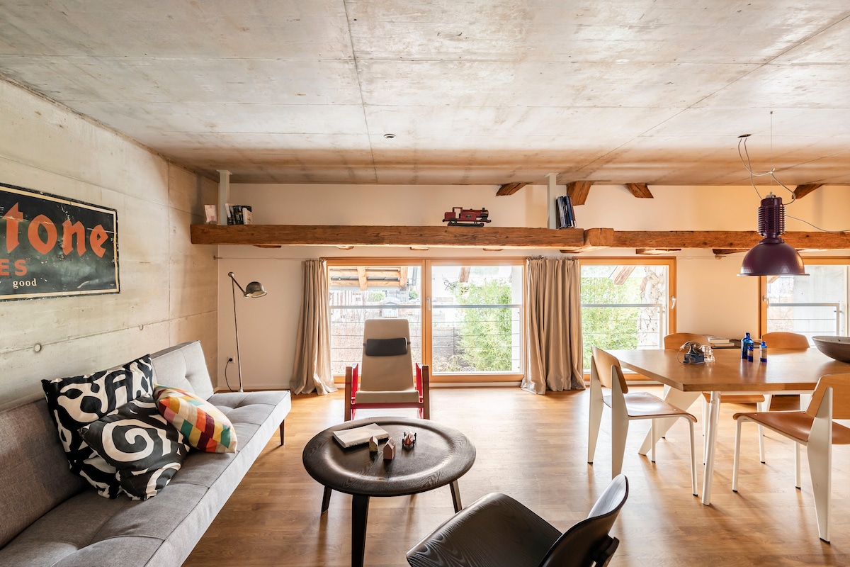 A modern living space features wooden beams and a concrete ceiling, complemented by large windows that bring in natural light. A comfortable sofa, decorated with colorful cushions, sits alongside a wooden dining table surrounded by chairs. A relaxed atmosphere is created by the mix of materials and furnishings.