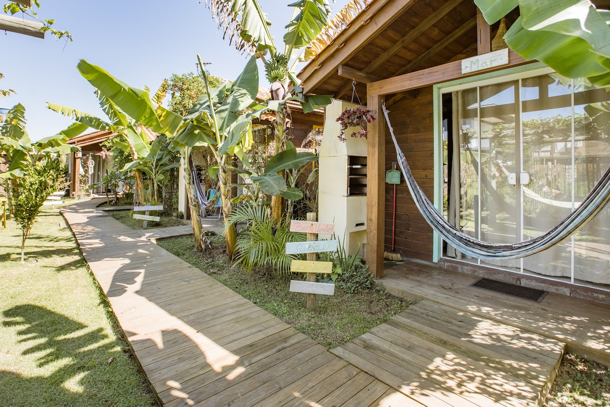 A pathway made of wooden planks extends between well-maintained gardens, bordered by tropical foliage. Several charming cabanas are visible, each featuring a small porch and a hammock, creating a relaxed outdoor atmosphere. Sunlight brightens the scene, contributing to the tranquil setting.