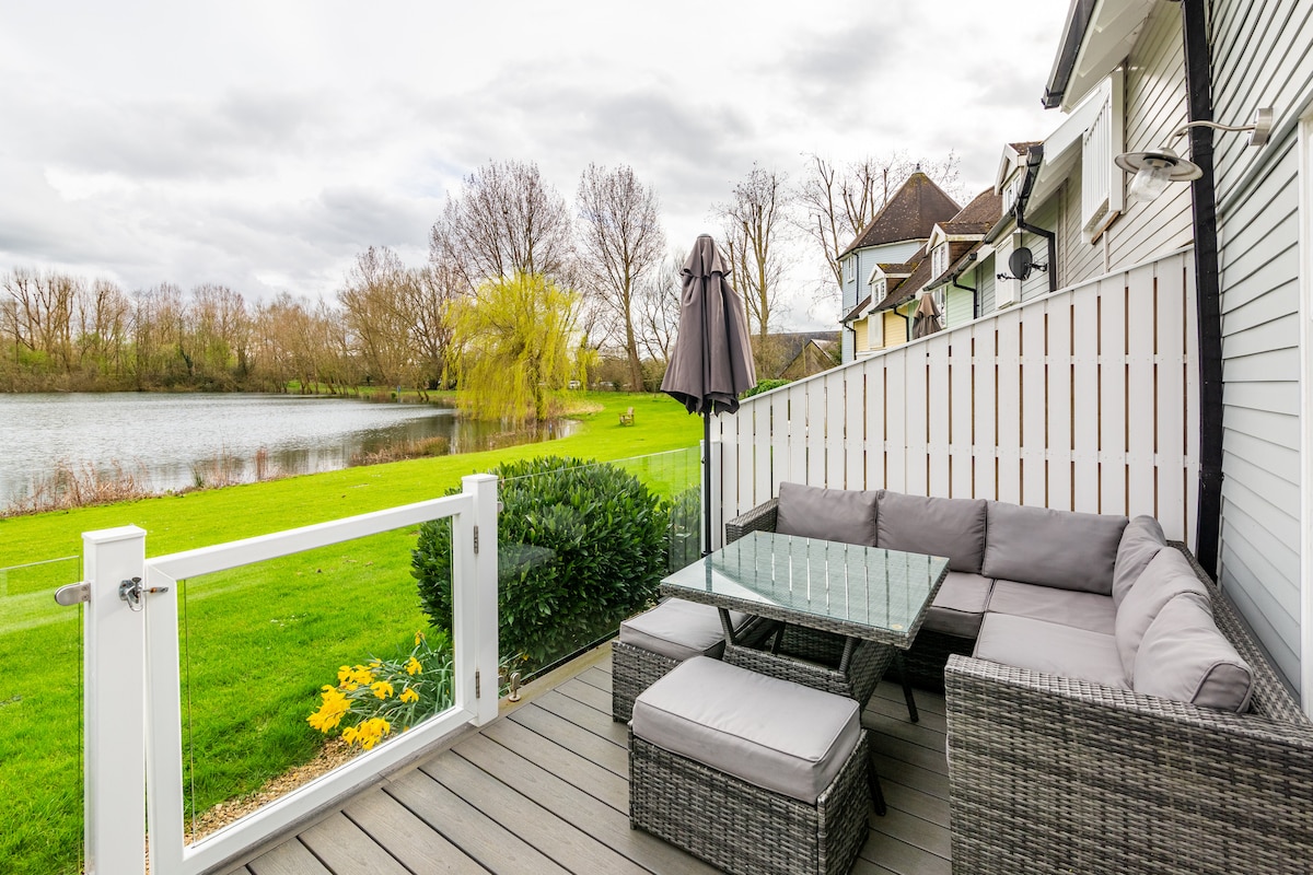 A spacious decking area features comfortable seating with a glass table, overlooking a serene lake. Lush green grass surrounds the space, while trees are visible along the water's edge. A patio umbrella stands nearby, ready for use on sunny days.