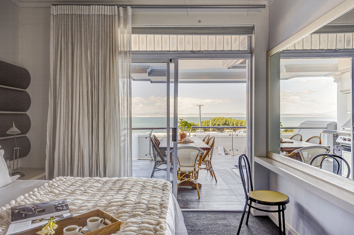 A serene bedroom is complemented by light-colored decor and a cozy knit blanket. A sliding glass door opens to a spacious patio with outdoor furniture and sweeping views of the sea. Natural light floods the room, reflecting off a stylish mirror.