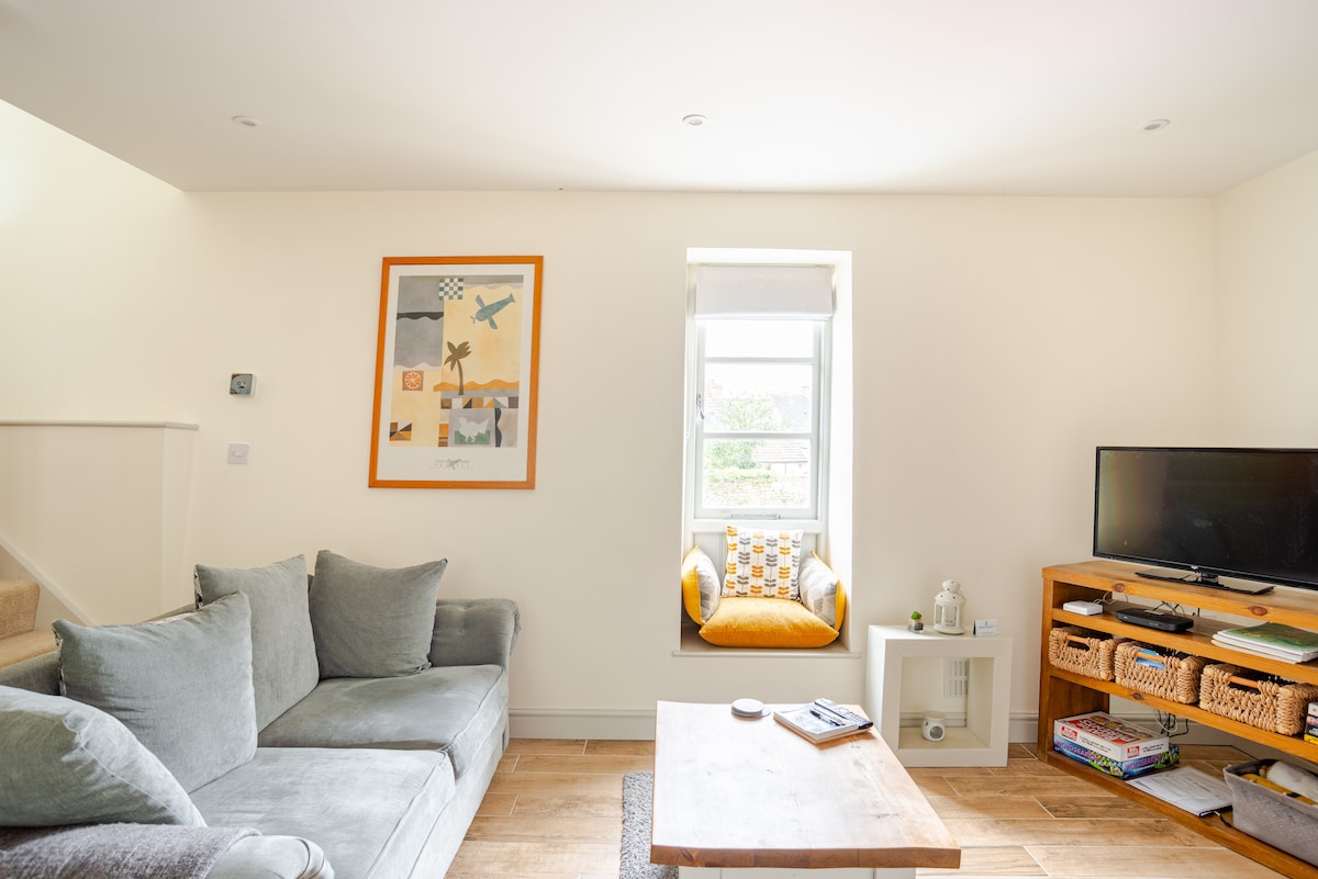 A bright living area features a comfortable gray sofa with multiple cushions. An art piece adorns the wall above, complementing the light ambiance. A small window nook with a yellow accent chair adds charm, while a TV and a neatly organized side table enhance the space.