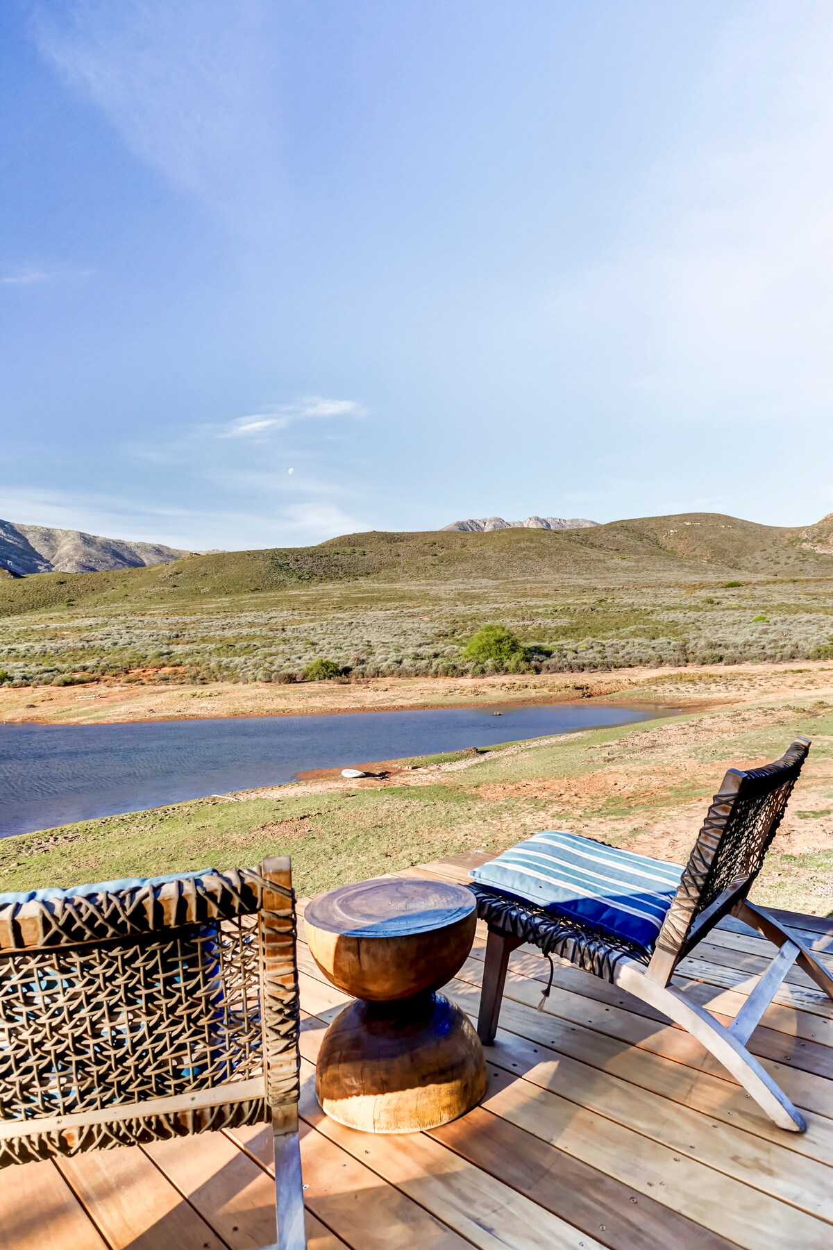 A wooden deck features two chairs and a round table, offering a view of the river and surrounding mountains. The landscape showcases green vegetation and a clear sky, enhancing the tranquil setting for relaxation.