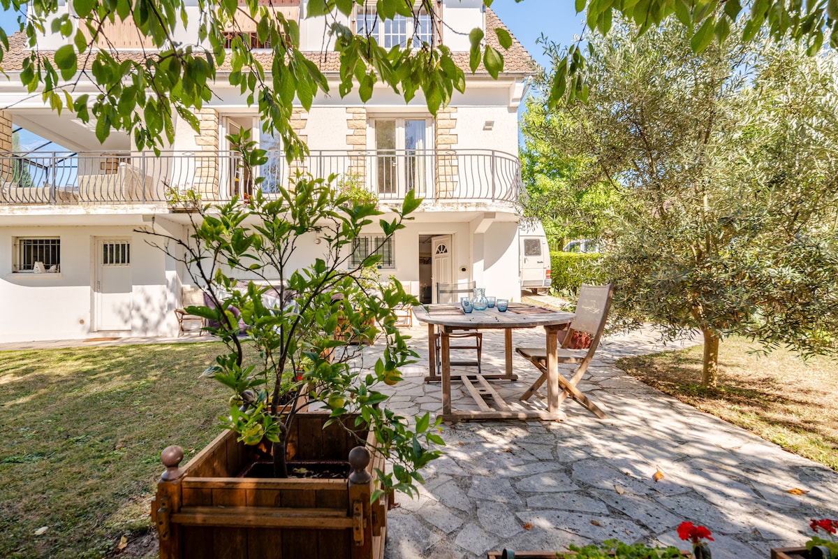 A spacious outdoor area is showcased, featuring a wooden dining table surrounded by chairs. Lush greenery, including trees and plants, is present, providing a serene environment. The house is visible in the background, with large glass doors leading to the interior, enhancing the connection to nature.