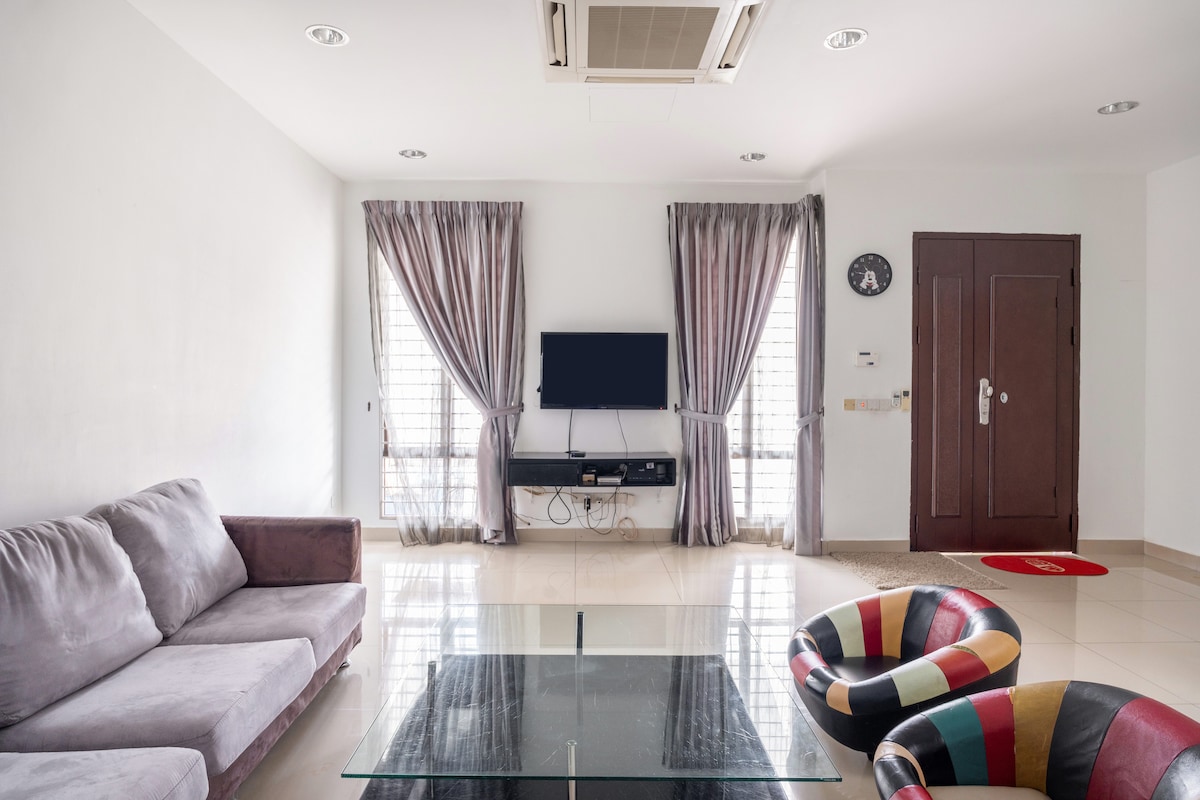 The living room features a comfortable sofa set and a coffee table with a glass surface. A television is mounted on the wall, positioned for easy viewing. Natural light is let in through two large windows dressed in light curtains, enhancing the room's spacious feel.