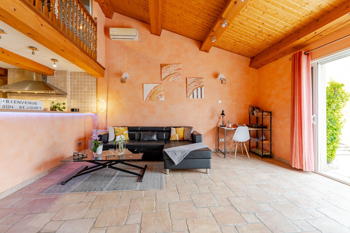 An inviting living area features a black sectional sofa and a coffee table, complemented by decorative artwork on peach-colored walls. A workspace with a desk and chair is present, and the room is illuminated by natural light from sliding glass doors leading to an outdoor space.