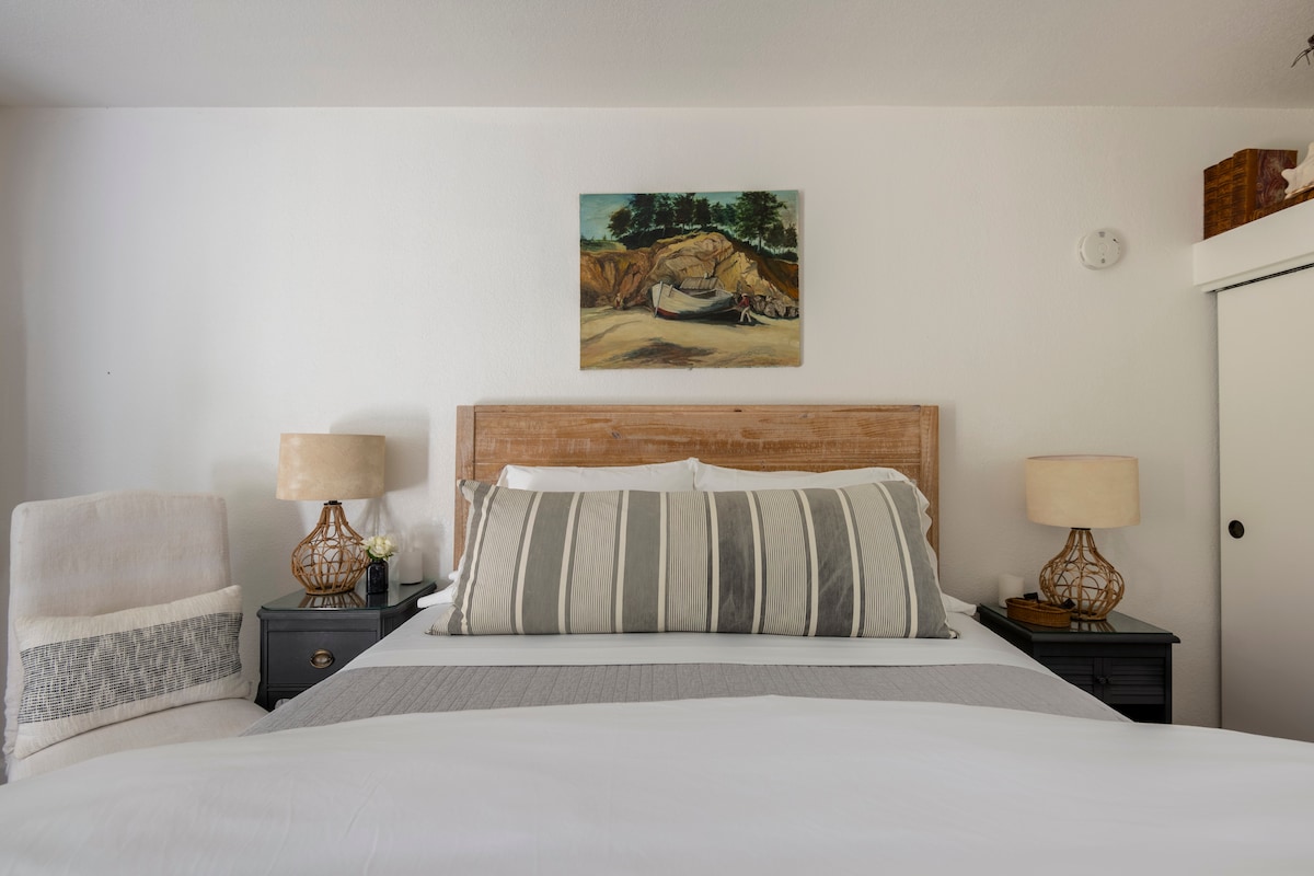 A cozy sleeping area features a queen bed, topped with striped pillows and a gray blanket. Nightstands with golden lamp bases are positioned on either side of the bed, and a painting of a serene landscape hangs above. Natural light brightens the space, complemented by neutral-colored walls.