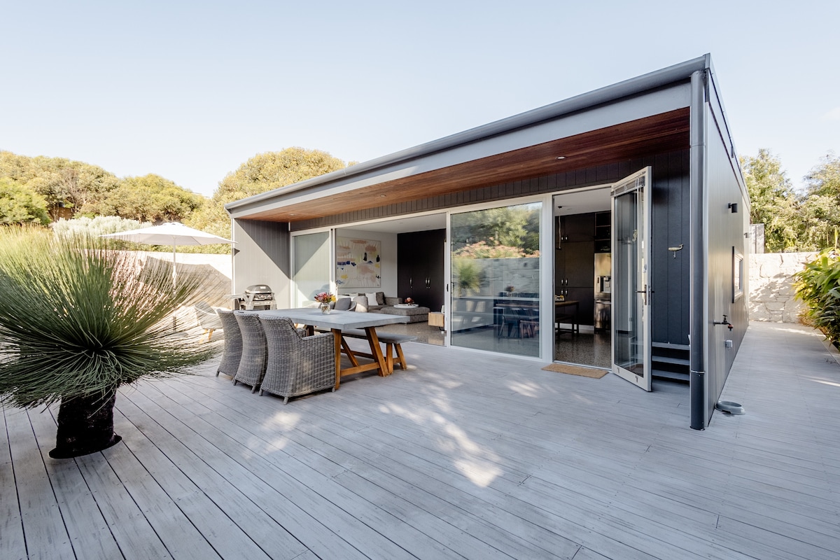 An outdoor dining area is visible on a spacious deck, featuring a wooden table surrounded by woven chairs. Large glass doors open to the interior, allowing natural light to flow in. Lush greenery and stylish decor enhance the inviting outdoor space.