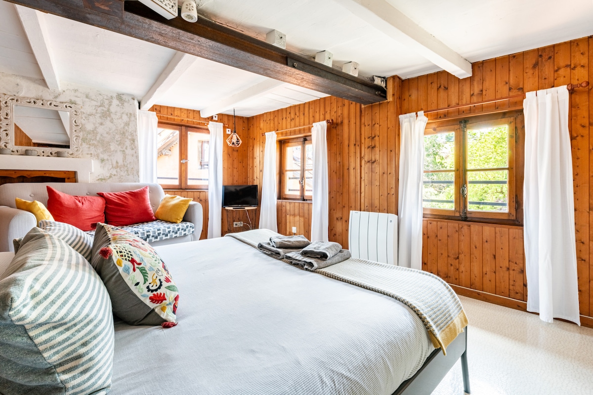 A warm, inviting bedroom is highlighted by wooden paneling and large windows, allowing natural light to illuminate the space. A spacious bed is adorned with decorative pillows and a soft throw, and a small TV is positioned for convenience. Two white curtains frame the windows.