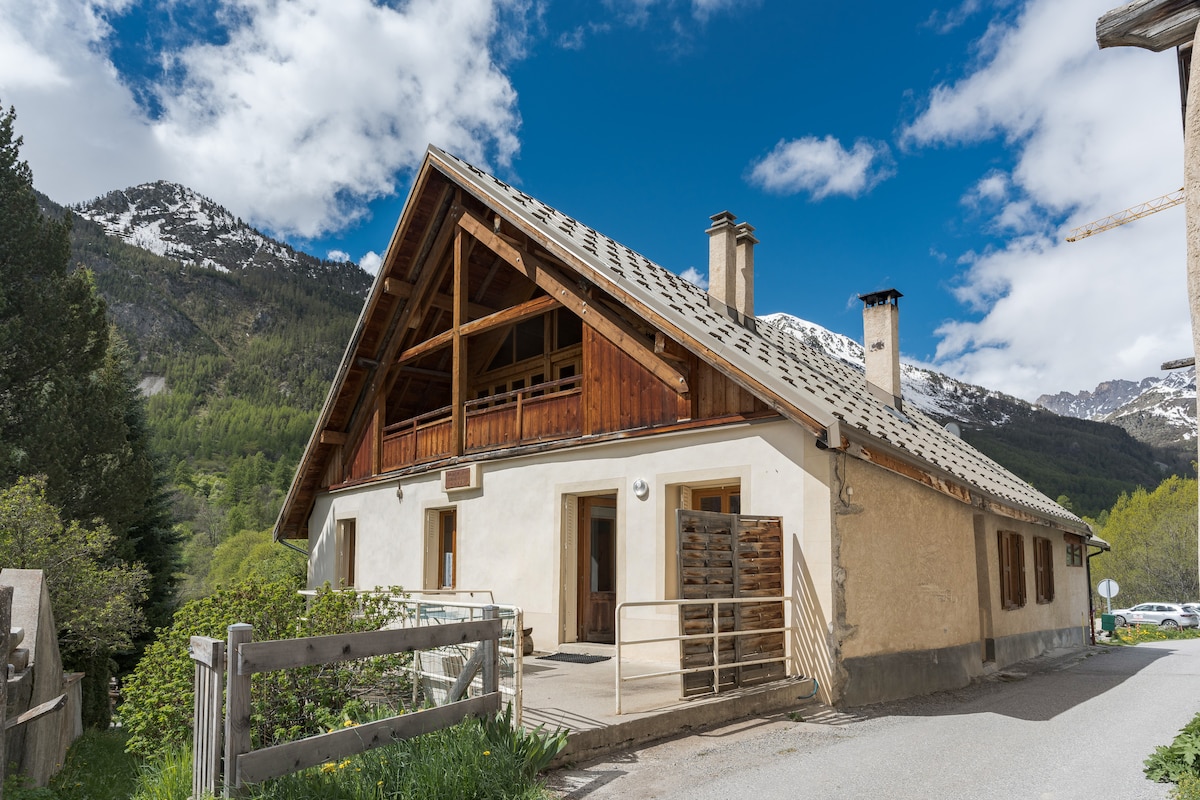 The exterior of a historic house is displayed, featuring a sloped wooden roof and large windows that allow natural light to enter. Surrounding mountains and greenery create a scenic backdrop, while a pathway leads toward the entrance, enhancing access to the property.