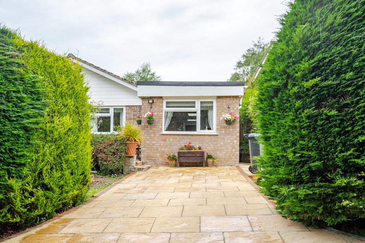 A welcoming entrance to the studio apartment is framed by lush hedges, creating a sense of privacy. The paved pathway leads to a well-lit front door adorned with potted flowers, inviting guests into the cosy space beyond.