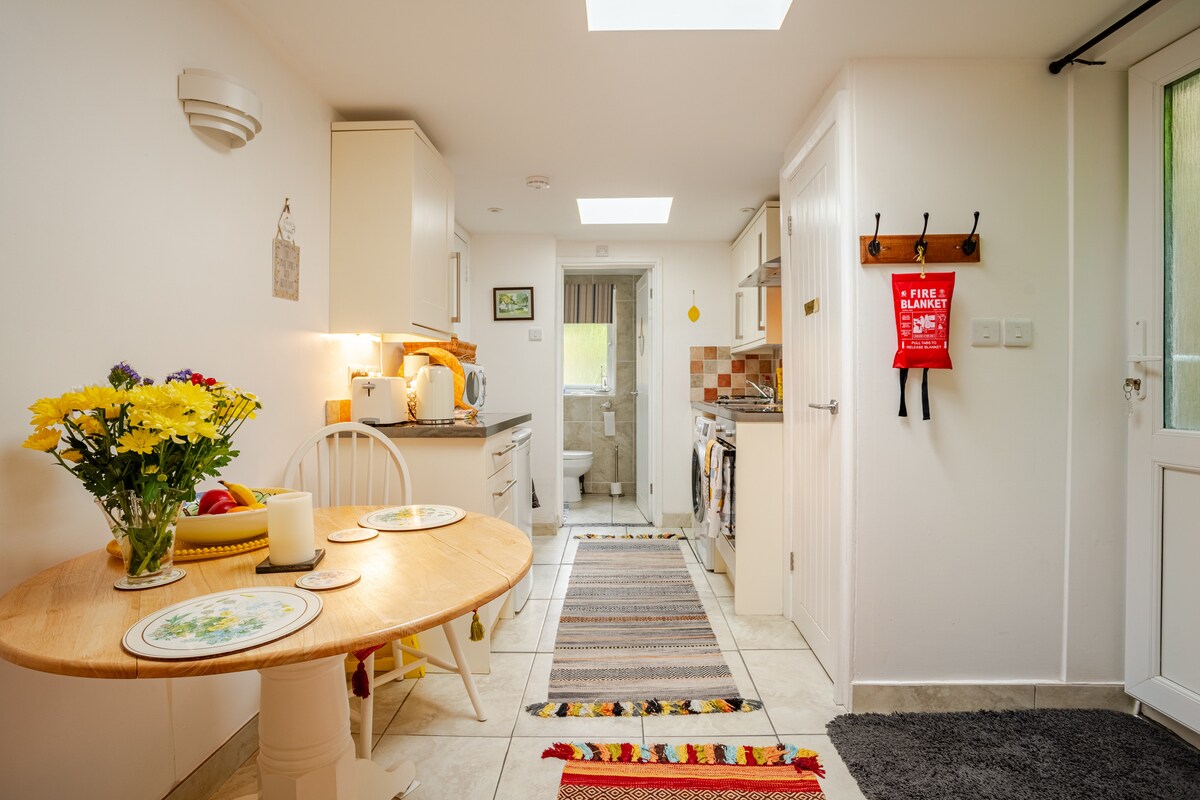 A bright kitchenette and dining area are showcased with a round wooden table set for two, accompanied by a vase of flowers. The modern kitchen features appliances, ample storage, and an area rug. Natural light enters through a skylight, illuminating the inviting space.