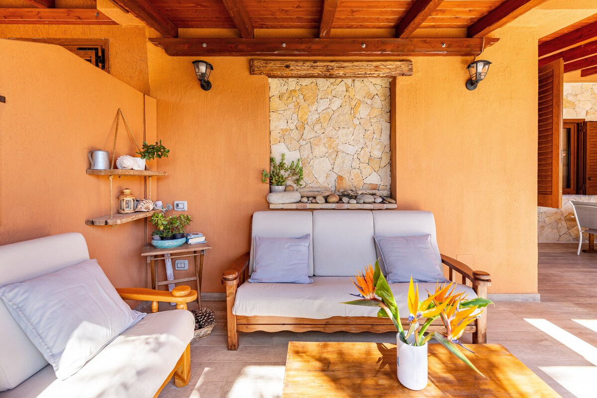 A comfortable seating area features a light-colored sofa and wooden chairs, complemented by a coffee table. Natural stone accents are visible in the wall behind, while a potted plant and decorative items add a touch of greenery.