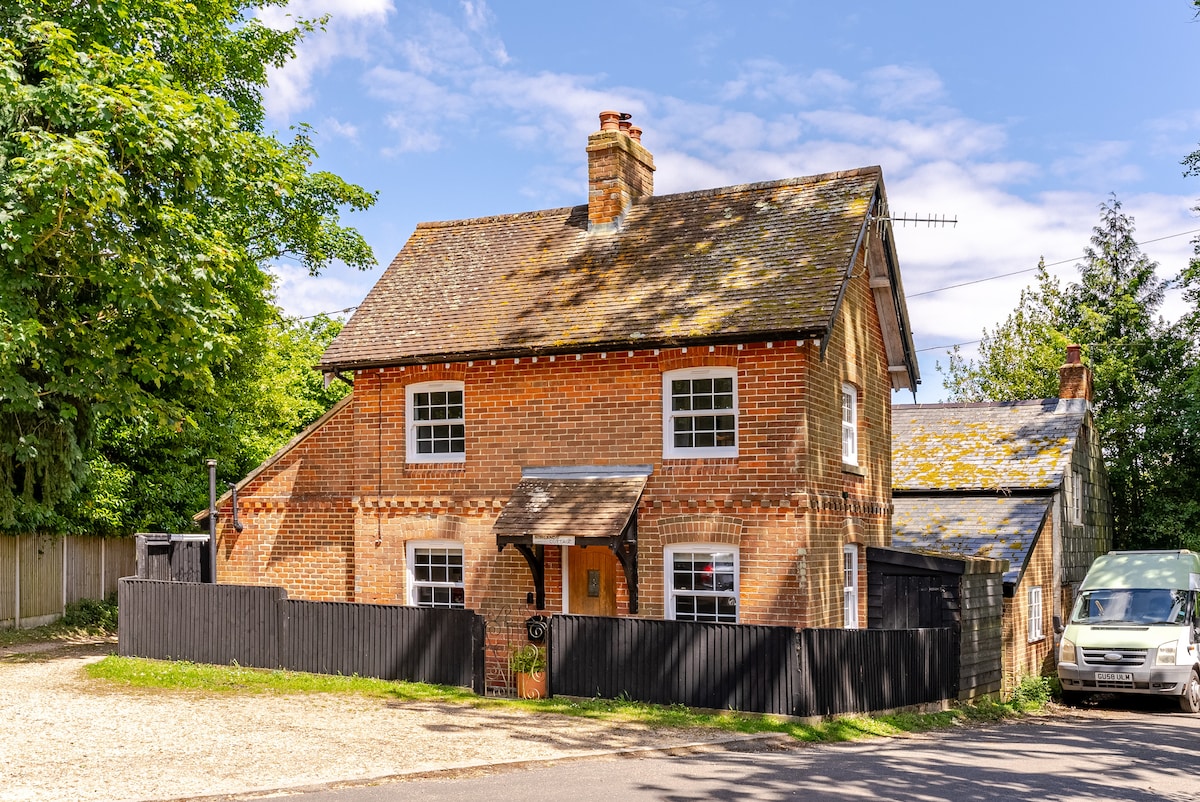 The cottage is a charming, newly refurbished brick structure with a sloped roof and a welcoming front entrance. A gravel area is visible in front, providing parking space, while a fenced yard surrounds the property. Lush greenery from nearby trees adds to the peaceful atmosphere.