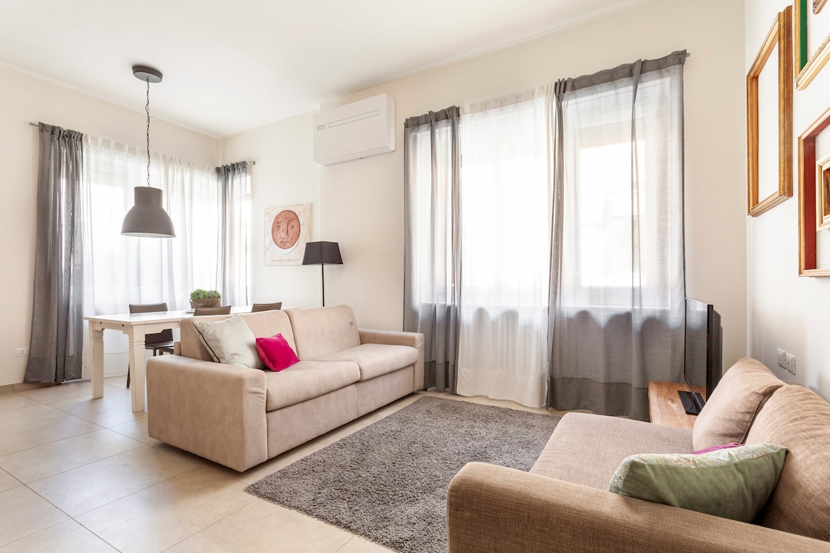 A light-filled living area features two sofas, one adorned with a pink accent pillow. A dining table is positioned nearby, surrounded by chairs, with large windows dressed in sheer curtains allowing ample natural light to illuminate the space.