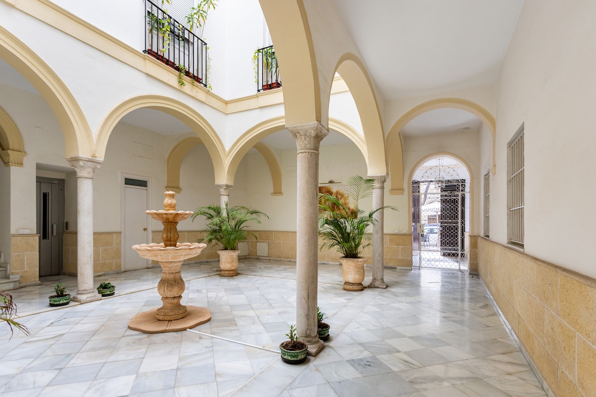 An indoor courtyard is surrounded by elegant arches, featuring a central fountain. Potted greenery is placed around the marble floor, enhancing the serene atmosphere. Light enters through open spaces, illuminating the intricate details of the architecture.