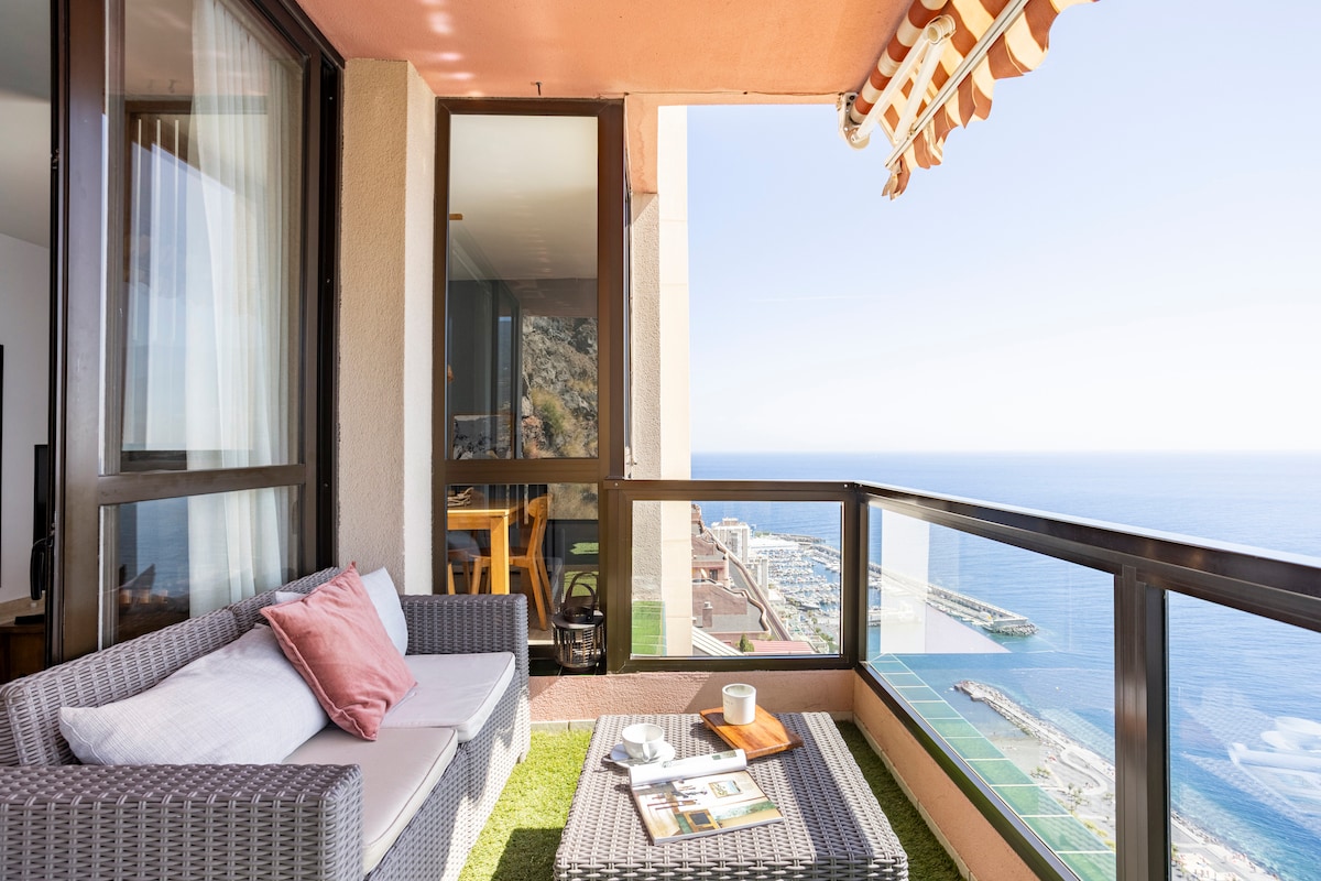A terrace equipped with outdoor seating offers panoramic views of the ocean. A small table is set with a cup and a small plate, while a soft blanket is draped over the seating area. The crisp blue sky complements the inviting atmosphere.