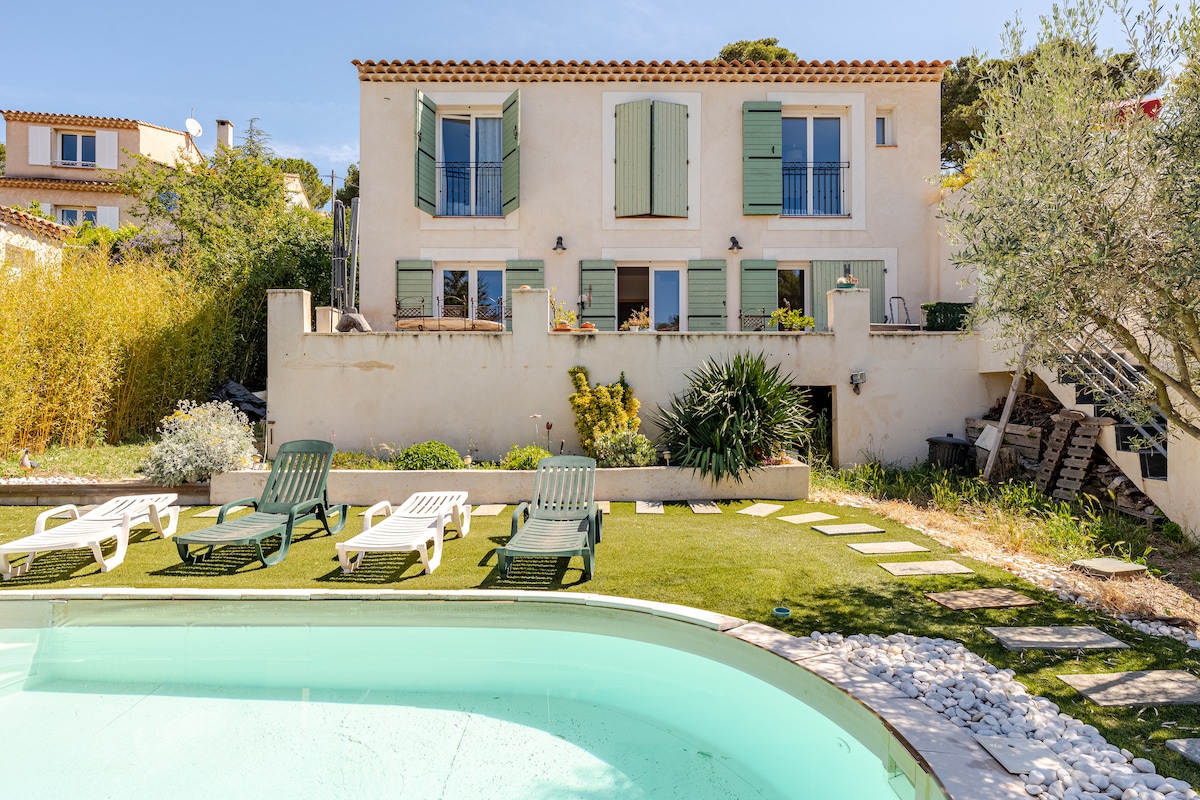 A charming villa is depicted with light green shutters and a variety of outdoor seating options arranged on a grassy area. A private pool is visible in the foreground, surrounded by smooth stones, and a stone pathway leads to the entrance of the home.