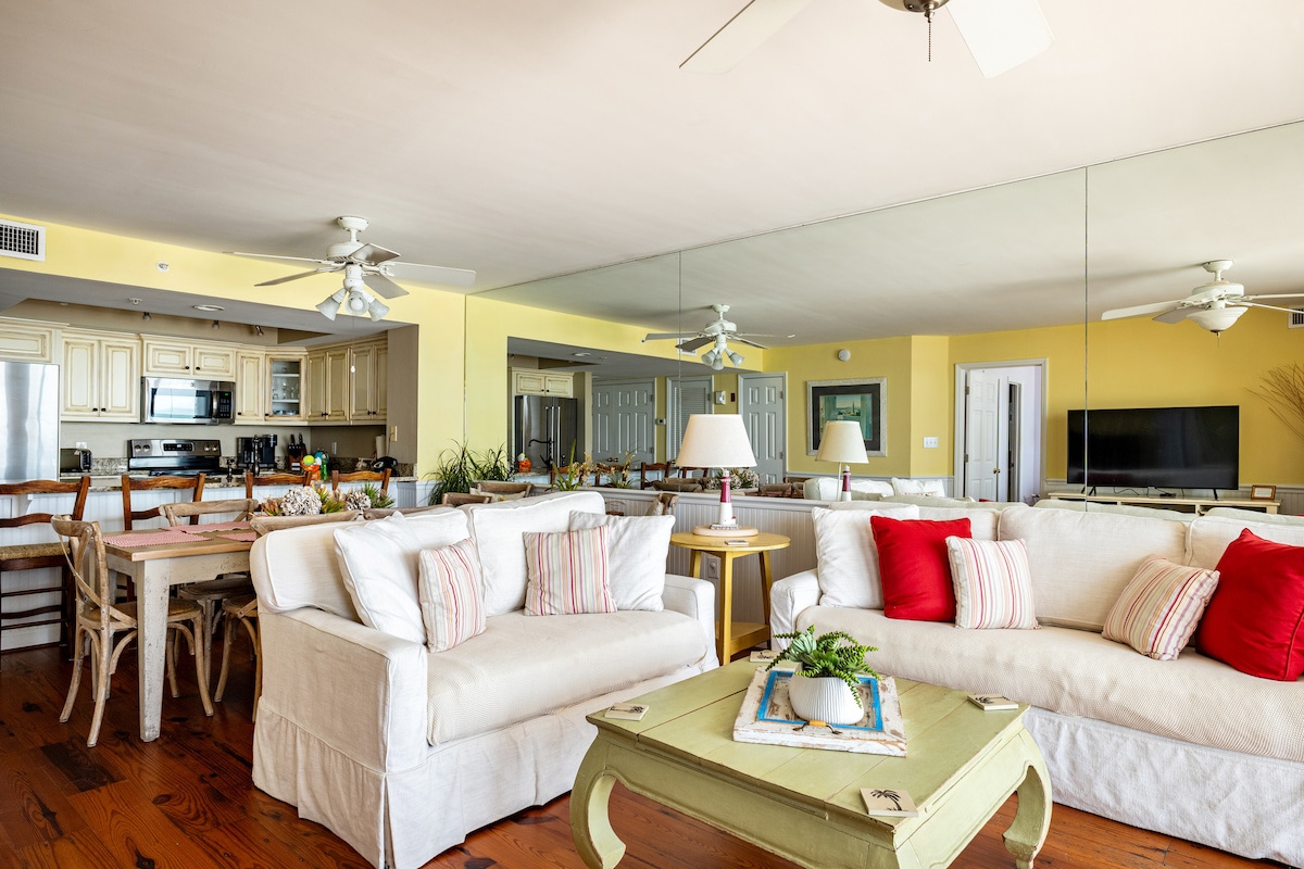 A spacious living area features comfortable sofas adorned with pillows, a coffee table, and a ceiling fan overhead. A large mirror reflects the brightness of the room. The open kitchen and dining area are visible in the background, showcasing a modern aesthetic and ample seating.