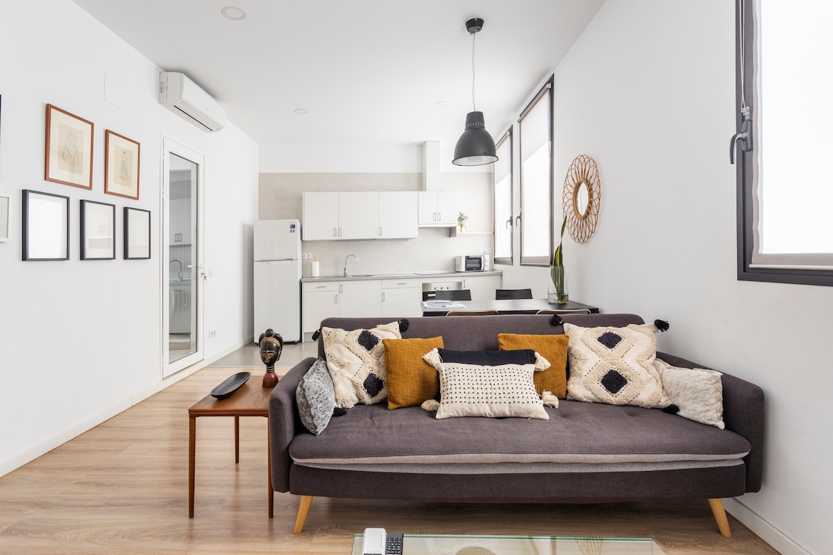 The living area features a modern sofa adorned with decorative cushions and a coffee table. A dining area with sleek furnishings is visible in the background, alongside an open kitchen equipped with appliances. Natural light floods the space through large windows.