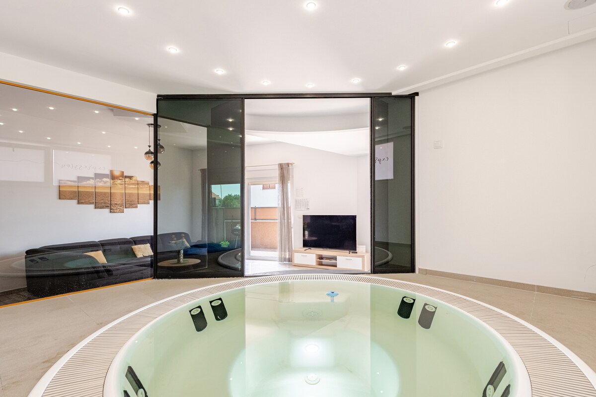 A spacious living area is visible, featuring a hot tub at the center. Large glass sliding doors allow natural light to fill the room. A comfortable sofa and a television are positioned in the background, creating an inviting space for relaxation.