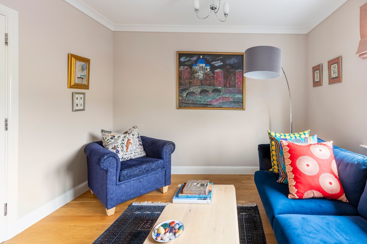 A lounge area is presented, featuring a blue sofa adorned with vibrant cushions and a matching armchair. A wooden coffee table is center stage, displaying decorative items. Art pieces are framed on the walls, and warm light is provided by a standing lamp.