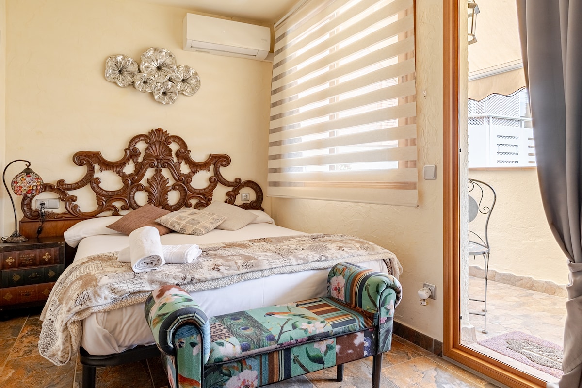 A sleeping area features a spacious bed with decorative pillows and a cozy blanket. An ornate wooden headboard adds character, while a colorful bench sits at the foot. Light filters through partially opened striped window blinds, creating a warm ambiance in the room.
