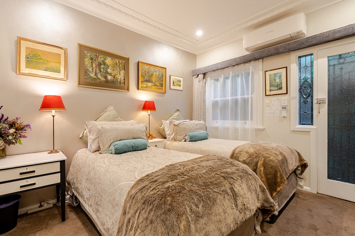 A well-lit room features two spacious beds adorned with soft throw blankets and decorative cushions. Art pieces hang on the pale walls, while bedside lamps provide a warm glow. Light filters through sheer curtains, contributing to a tranquil ambiance. A private entrance is visible in the background.