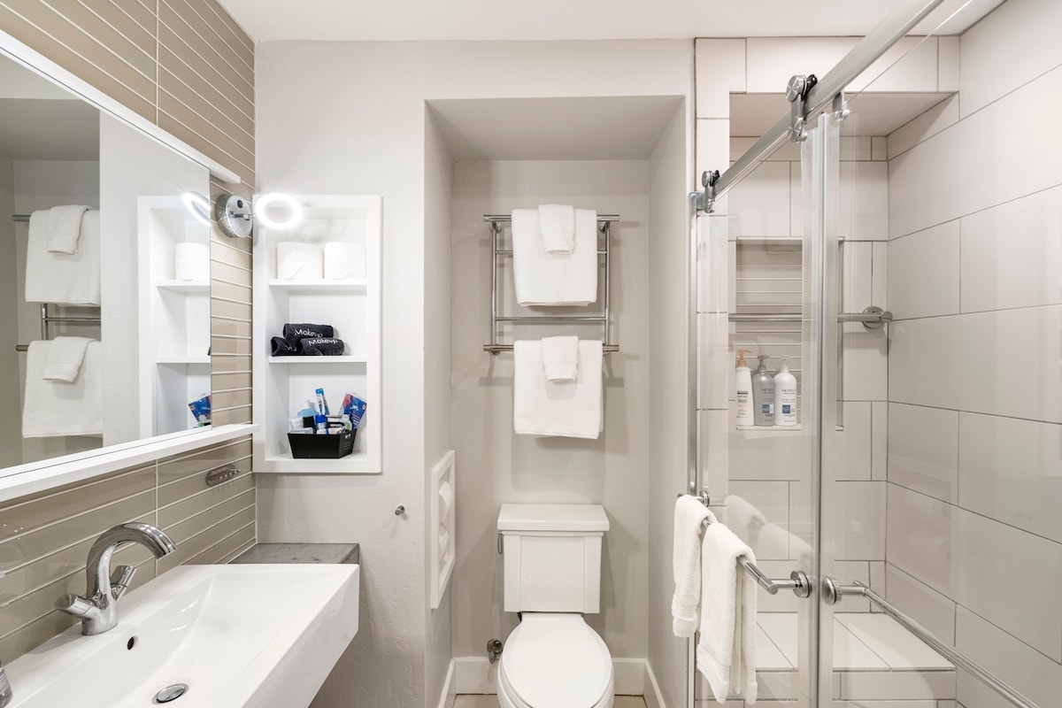 The bright bathroom features a walk-in shower with glass doors, equipped with toiletries and plush towels on a chrome rack. A large mirror above a sleek sink complements the clean, modern tiles, enhancing the overall spacious feel.