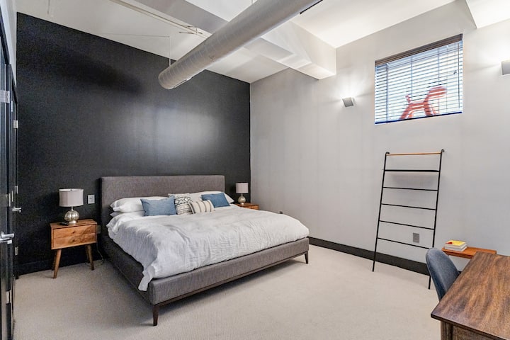 A King Bedroom with premium Casper mattress sits on an upholstered Restoration Hardware bed frame and flanked by two side tables with reading lamps. Behind a window with wood blinds sits a Herman Miller Magis Puppy who keeps watch of the sidewalk.