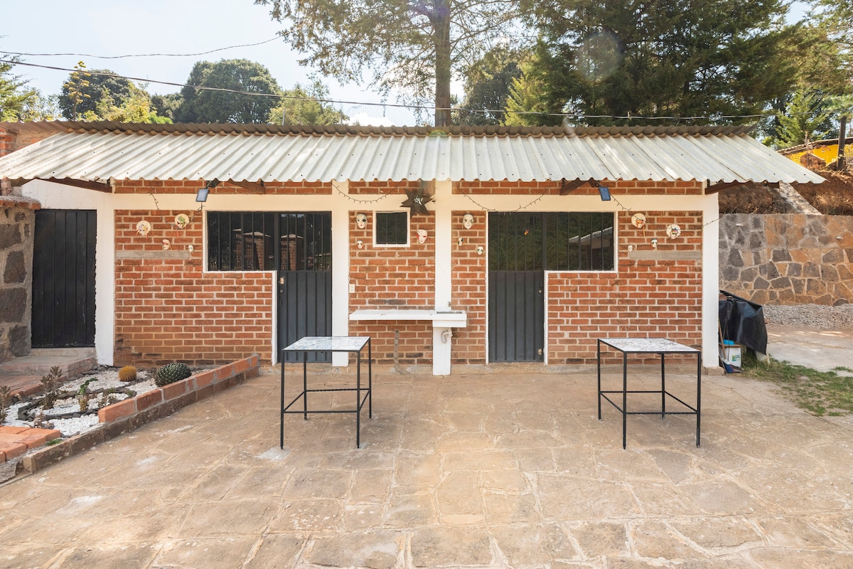 An exterior view of a brick cabin is shown, featuring two entrances framed by dark doors. Two metal tables are situated on the stone patio, surrounded by a neutral-toned landscape with minimal vegetation. A canopy provides coverage above.