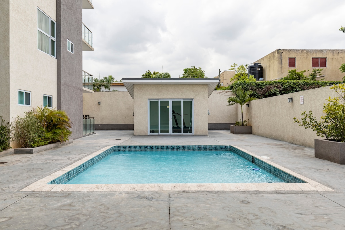 A modern pool area is depicted, featuring a rectangular swimming pool surrounded by a spacious concrete deck. Several green plants line the edges, and a glass-enclosed room is visible in the background, providing an open view of the pool.