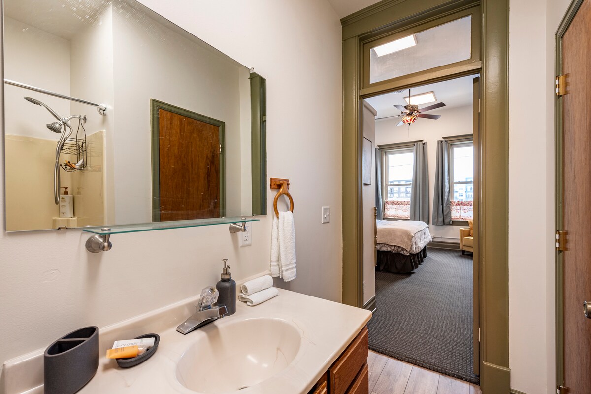 A bathroom is displayed featuring a modern sink and a large mirror reflecting the well-lit space. The doorway leads to a spacious bedroom beyond, where natural light enters through two windows adorned with patterned curtains.