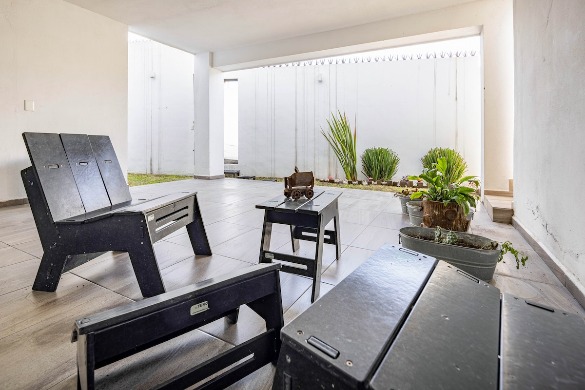A shaded terrace area features minimalistic wooden furniture, including a pair of chairs and a small table. Potted plants are arranged along the walls, and natural light brightens the space through openings above, creating a calm and inviting environment.