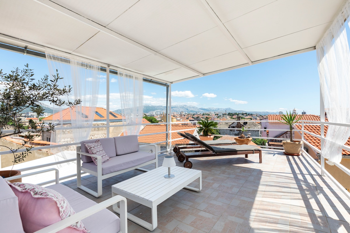 A covered terrace offers seating with a white sofa and lounge chair, complemented by a small table. Soft white curtains frame the space, enhancing the view of neighboring rooftops and green palm trees beneath a clear blue sky.