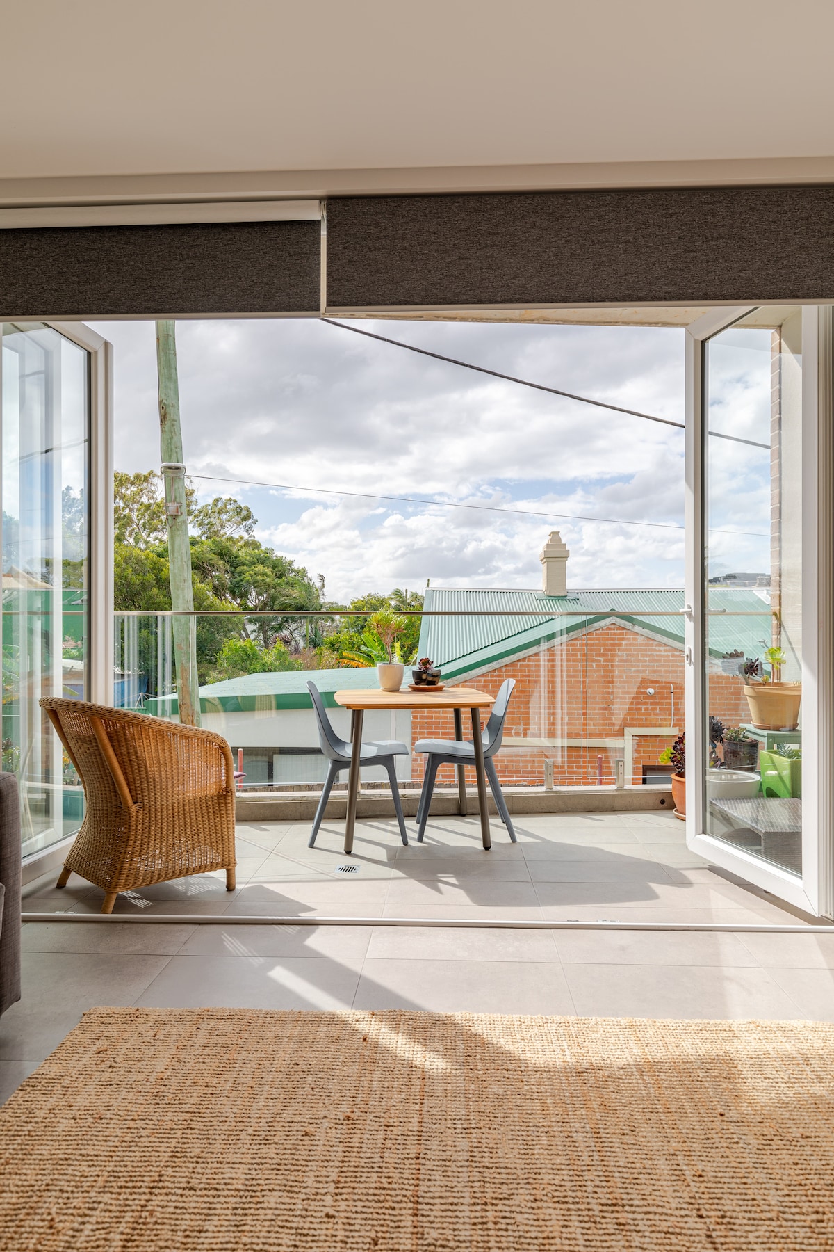 A covered balcony features a small round table with two chairs, surrounded by greenery and rooftops. Stackable glass doors open to the view, allowing natural light to fill the space. A textured rug lies on the floor, creating a cozy atmosphere.