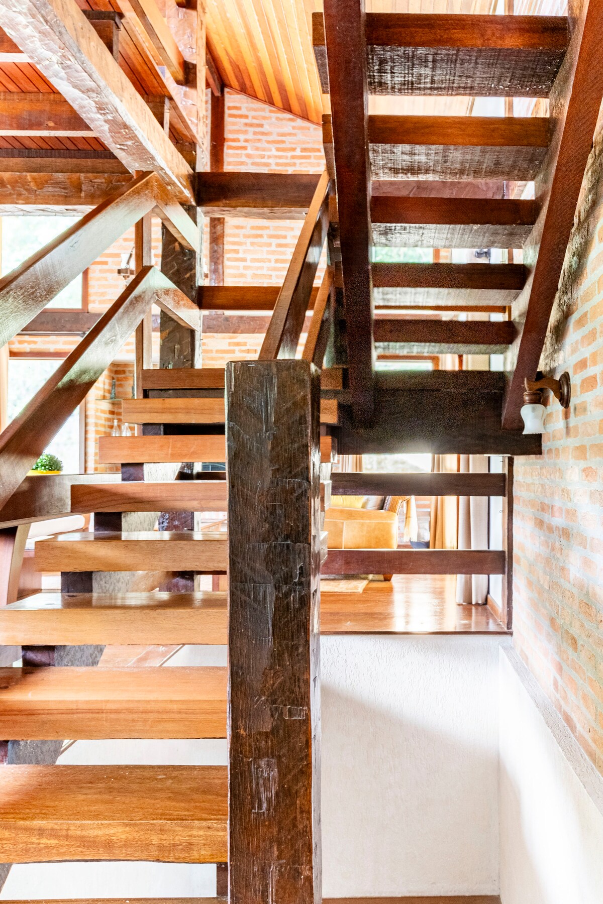 A wooden staircase is showcased, featuring open steps and a strong handrail. Natural light highlights the warm tones of the wood and the surrounding brick walls, creating a welcoming entryway. The staircase leads to an upper level, emphasizing the multi-level design of the space.