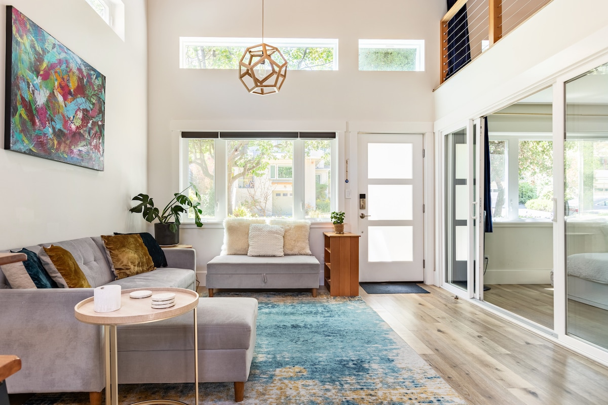 A bright and open living area is highlighted by large windows allowing natural light to flood in. A comfortable gray sofa and a matching daybed are positioned on a soft area rug. A small coffee table sits among decorative accents, and a door leads to the outside.