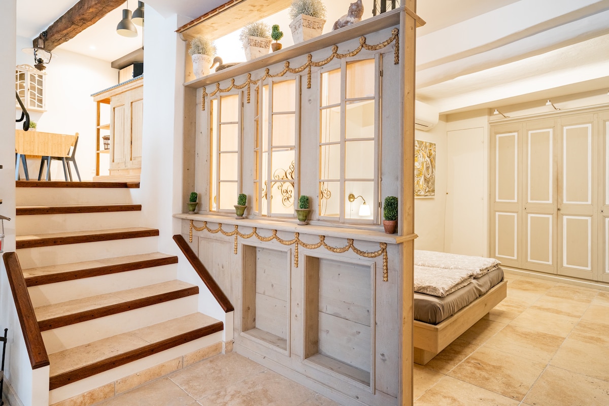 A wooden staircase leads to a semi-open space featuring a decorative console with glass-fronted cabinets. Soft neutral tones and natural finishes create a harmonious ambiance. A spacious bed is visible in the adjoining area, complemented by built-in closet space on the side.