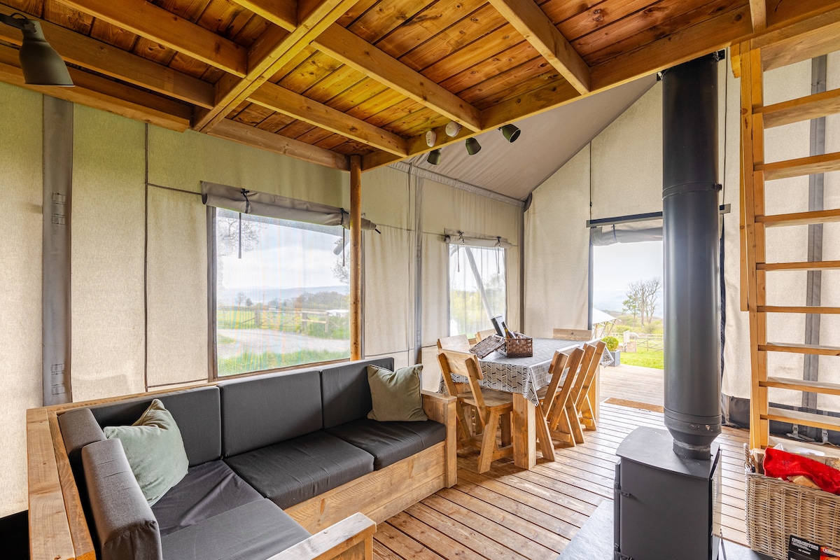 An open living area is depicted with wooden ceiling beams and large windows allowing natural light. A seating area features a wooden sofa with cushions, while a dining table with chairs is set near the window. A wood-burning stove adds warmth to the space.