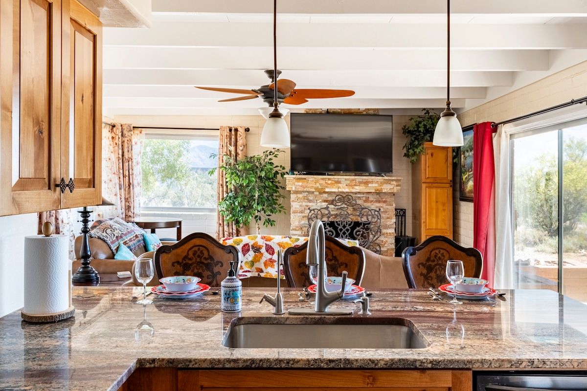 A kitchen countertop offers a clear view of a dining area, featuring a wooden dining table set for four. The space is enhanced by a comfortable living area with a stone fireplace and a large television. Natural light streams through the sliding glass doors, revealing desert views.