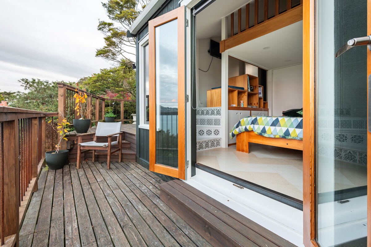 A deck area extends from the doorway, featuring two lounging chairs and planters. Brightly colored bedding is visible on a spacious bed inside, while the indoor space showcases wood and tile accents. Large glass doors provide an open view of the surrounding natural beauty.
