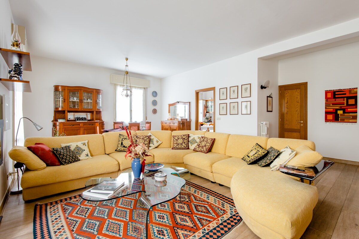 A spacious living area features a large yellow sectional sofa accented with patterned cushions. A round glass coffee table sits in the center, surrounded by a decorative area rug. Shelves display various decorative items, and sunlight streams in through large windows.