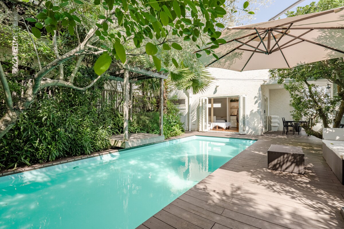 An outdoor swimming pool is set amidst lush greenery, with a wooden deck surrounding it. An umbrella provides shade, and glass doors lead into the interior space. The tranquil water reflects the natural surroundings, creating a serene atmosphere.