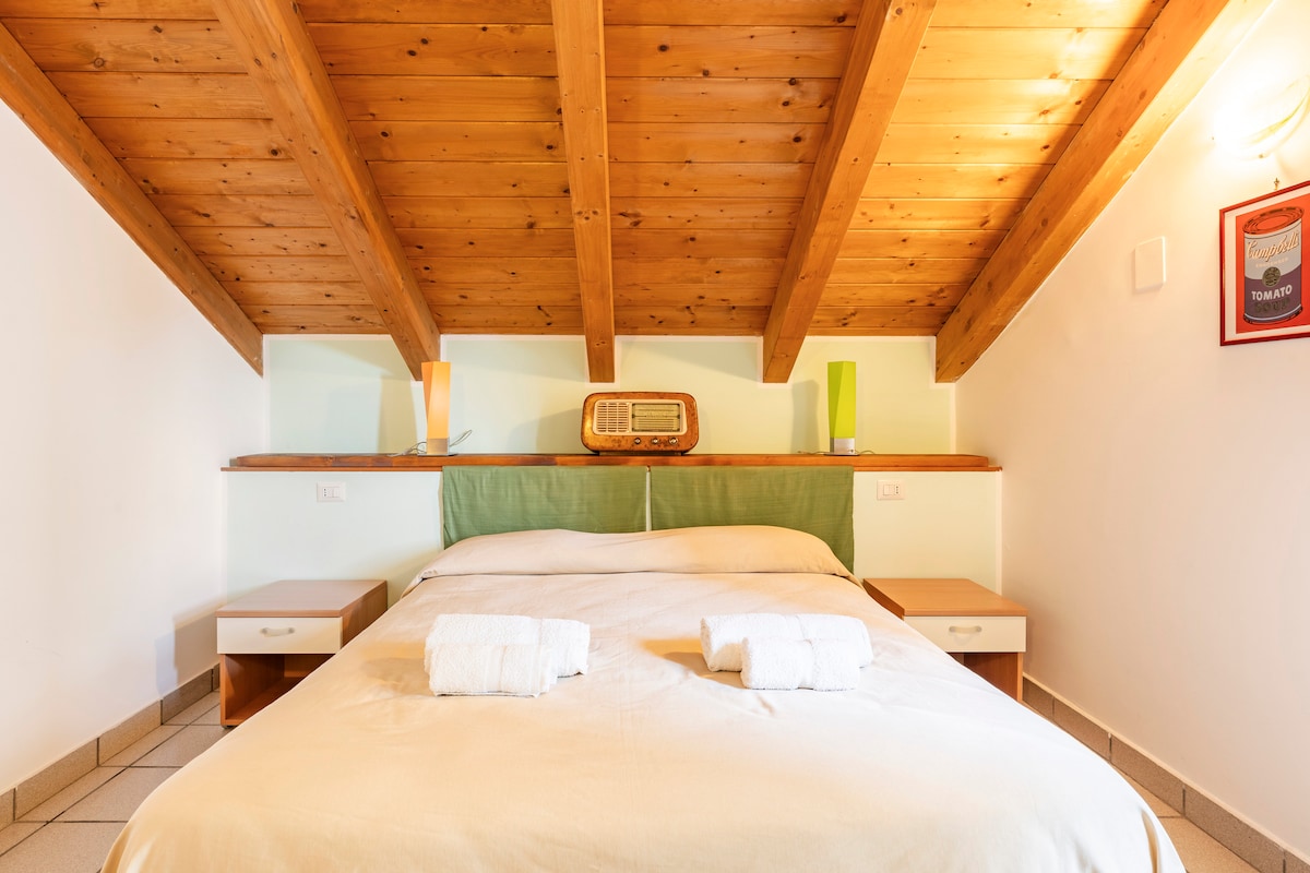 A spacious sleeping area features a double bed with a light-colored blanket and two white towels neatly placed on it. The wooden sloped ceiling adds warmth, while green accents are highlighted on the headboard. Two bedside lamps provide soft illumination.