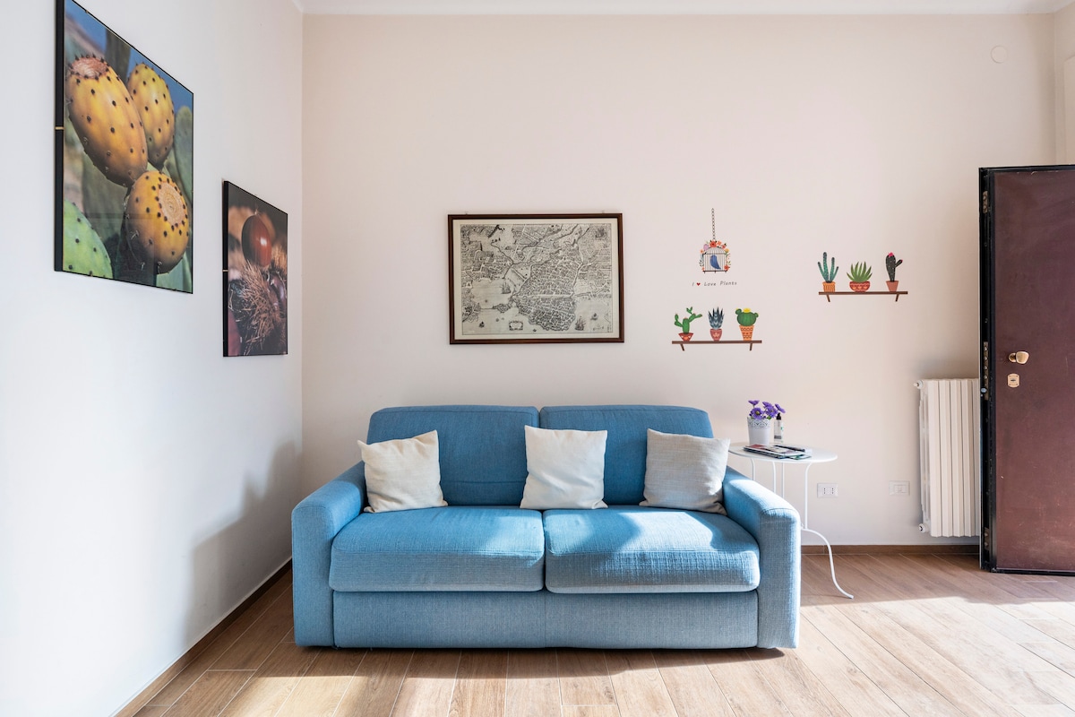 A light blue sofa is positioned against a neutral wall, paired with two white cushions. Colorful wall art, including framed photographs and illustrated plants, decorates the space. A small table with a vase of purple flowers is nearby, and natural light illuminates the wooden flooring.