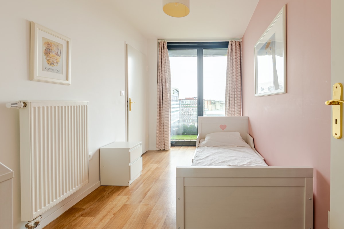 A bright, minimalistic bedroom features a single bed with a heart-shaped detail. A nightstand and a radiator are present. Large glass doors lead to a balcony, allowing natural light to fill the space, while light curtains softly frame the windows.