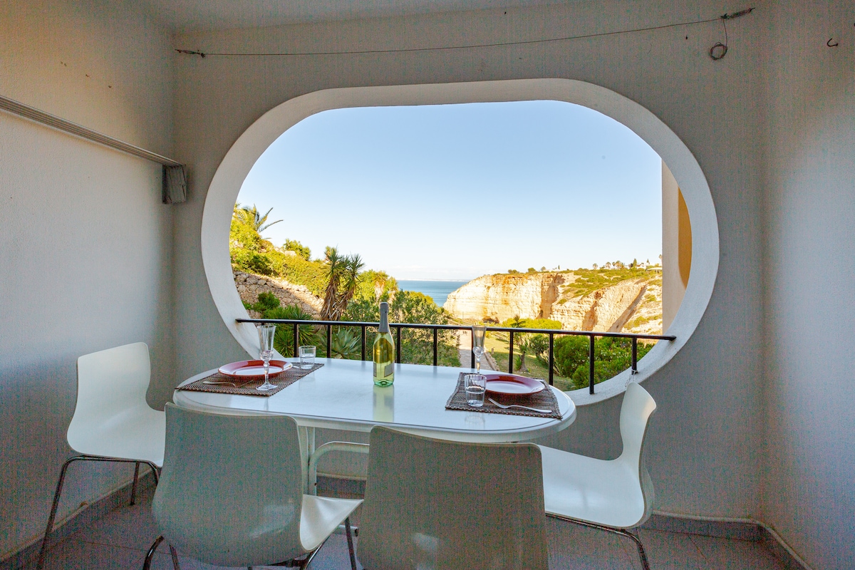 A dining area is visible, featuring a round table with white chairs. A large oval window frames a scenic view of the coastline, allowing natural light to fill the space. The setting is designed for enjoying meals while taking in the landscape.