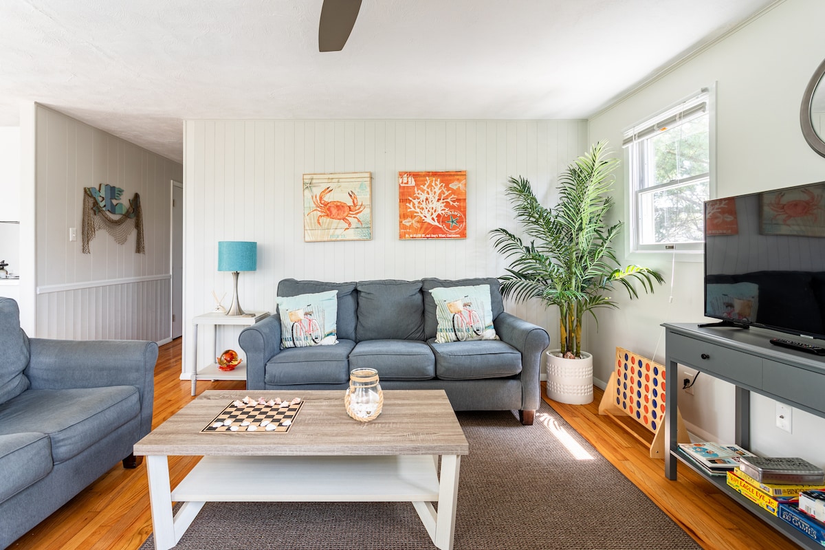 A living area is presented with soft blue sofas anchored around a central coffee table. Bright artwork featuring crabs is displayed on the walls. A large indoor plant adds a touch of greenery, while natural light streams in through the window, enhancing the relaxed ambiance.